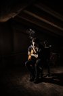 Парень играет на гитаре и курит сигарету на стуле на чердаке в темноте — стоковое фото