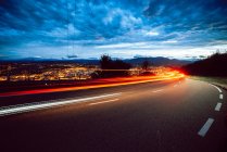 View of bright trail lights on route in evening in countryside on city background and cloudy sky — Stock Photo