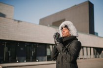 Stylish African American male in fur hat standing against modern building on sunny day and rubbing hands — Stock Photo