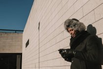 Homme afro-américain en chapeau de fourrure appuyé sur un mur de briques avec téléphone portable sur une journée ensoleillée dans la rue de la ville — Photo de stock
