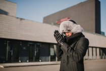 Stylish African American male in fur hat standing against modern building on sunny day and rubbing hands — Stock Photo