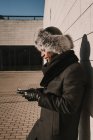 Attraente uomo afroamericano in cappello di pelliccia appoggiato sul muro di mattoni con il telefono cellulare nella giornata di sole sulla strada della città — Foto stock