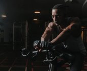 Homem Africano Americano confiante sentado em bicicleta de exercício durante o treino no ginásio escuro — Fotografia de Stock