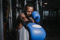 Homme noir en gants de boxe bleus frappant l'air tout en travaillant dans la salle de gym moderne sombre — Photo de stock