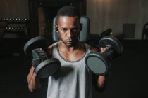 Young African American man lifting two heavy dumbbells and listening to music during training in modern gym — Stock Photo