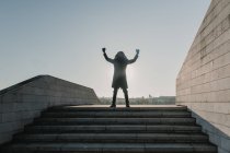 Афроамериканец в теплой одежде смотрит вверх и жестикулирует руками, празднуя победу на вершине лестницы в солнечный день в городе — стоковое фото