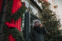 African American man in warm clothes talking on smartphone while standing on city street near building and conifer tree decorated for Christmas — Stock Photo