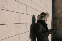 Attractive African American man in fur hat leaning on brick wall with mobile phone on sunny day on city street — Stock Photo