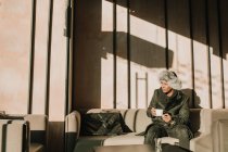Bell'uomo afroamericano in abito caldo che tiene una tazza di bevanda calda e distoglie lo sguardo mentre si siede su un comodo divano nel caffè — Foto stock