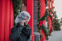 African American man in warm clothes talking on smartphone while standing on city street near building and conifer tree decorated for Christmas — Stock Photo