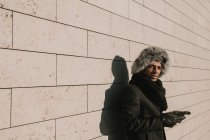 Привлекательный афроамериканец в меховой шапке, опирающийся на кирпичную стену с мобильным телефоном в солнечный день на городской улице — стоковое фото