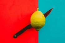 Sharp knife cutting juicy ripe lemon on vibrant red and blue background — Stock Photo