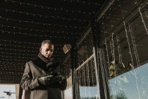 Afrikanisch-amerikanischer Mann mit modernem Smartphone, während er in der Nähe moderner Gebäude steht und Weihnachten mit Lichtergirlanden dekoriert — Stockfoto