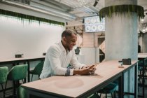 Side view of handsome African American man smiling and using modern smartphone while sitting at table in stylish cafe — Stock Photo
