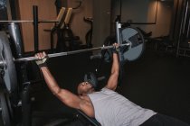 Hombre afroamericano guapo en ropa deportiva acostado en el banco y levantando pesadas pesas durante el entrenamiento en el gimnasio moderno - foto de stock