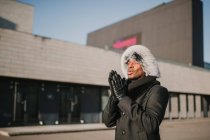 Привлекательный афроамериканец в меховой шапке стоит возле современного здания в солнечный день на городской улице, массируя руки — стоковое фото