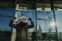 Handsome African American man in warm clothes smiling and gesturing with hands while celebrating success near glass wall of modern building — Stock Photo