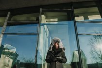 Черный человек в теплой одежде стоит у стеклянной стены современного здания и просматривает смартфон в солнечный день на городской улице — стоковое фото