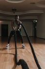 Нахабний афроамериканець, який виконує вправи з мотузками, працюючи в багатолюдній кімнаті сучасного спортзалу. — стокове фото