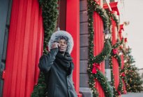 Афроамериканец в теплой одежде разговаривает по смартфону, стоя на городской улице рядом со зданием и хвойной елкой, украшенной на Рождество — стоковое фото