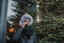 Афроамериканець у теплому одязі розмовляє на смартфоні, стоячи на вулиці біля будинку і хвойного дерева, прикрашеного Різдвом. — стокове фото
