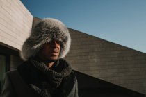 Attractive African American man in fur hat standing near a modern building on sunny day on city street — Stock Photo