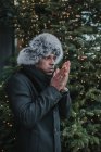 Handsome African American man in warm clothes rubbing hands while standing near Christmas tree with fairy lights on city street — Stock Photo