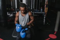 Beau mâle noir assis sur la machine d'exercice et portant des gants de boxe bleus pendant l'entraînement dans la salle de gym moderne — Photo de stock