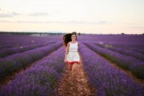 Feliz dama atractiva en vestido corriendo entre filas de hermosas flores púrpuras en el campo de lavanda - foto de stock