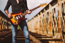Erwachsener Mann mit E-Gitarre steht an sonnigem Tag im Grünen auf verwitterter Brücke — Stockfoto