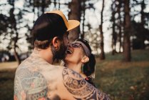 Вид сзади на молодого мужчину без рубашки в татуировках со снэпбеком обнимающего женщину в парке на размытом фоне — стоковое фото
