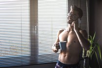 Uomo senza maglietta in piedi vicino a una finestra tende parlare sul telefono cellulare e in possesso di una tazza di caffè — Foto stock