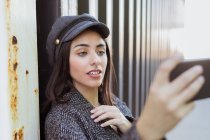 Очаровательная латиноамериканка в пальто и кепке делает селфи возле металлической стены — стоковое фото