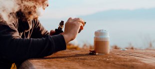 Persona irriconoscibile che vaporizza e utilizza lo smartphone per scattare foto di una tazza di caffè fresco seduto a un tavolo di legno in campagna — Foto stock