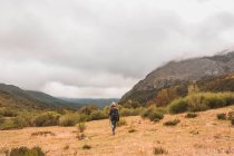 Vue arrière de dame en chapeau et veste de ski avec sac à dos et bâton de marche marchant sur la prairie près de la montagne dans les nuages à Isoba, Castille et Léon, Espagne — Photo de stock