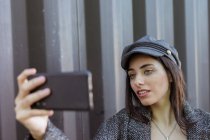 Charmante hispanische Dame in Mantel und Mütze macht Selfie in der Nähe der Metallwand — Stockfoto
