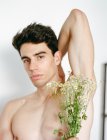 Vista lateral do jovem sem camisa com flores brancas frescas nas mãos olhando para a câmera no fundo borrado — Fotografia de Stock