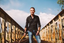 Adulto cara com guitarra elétrica em pé na ponte weathered e olhando para a câmera no dia ensolarado no campo — Fotografia de Stock