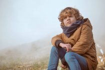 Adorabile ragazzo in abiti caldi guardando altrove mentre seduto sullo sfondo della maestosa collina nella giornata nebbiosa in campagna — Foto stock
