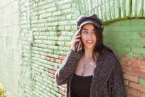 Souriant Hispanique jeune femme parlant sur téléphone portable près de mur de briques vertes — Photo de stock