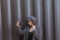 Стильная молодая женщина в пальто и кепке позирует и делает селфи возле металлической стены — стоковое фото