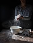 Crop lady battendo le mani nella farina vicino ciotola, scatola di uova e sbattere sul tavolo di legno su sfondo nero — Foto stock