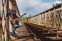 Erwachsener Mann mit E-Gitarre steht auf verwitterter Brücke und blickt an sonnigen Tagen in der Natur in die Kamera — Stockfoto