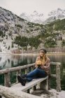Молода пані сидить на лавці заплющених очей біля дивовижної поверхні води між високими горами з деревами у снігу в Піренеях. — стокове фото