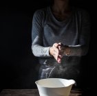 Crop lady clapping hands in flour near bowl, box of eggs and whisk on wooden table on black background — Stock Photo