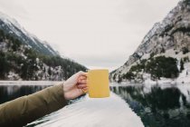 Coltivazione mano di azienda umana tazza gialla vicino incredibile vista della superficie dell'acqua tra alte montagne con alberi nella neve e cielo nuvoloso nei Pirenei — Foto stock