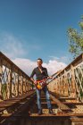 Adulto cara com guitarra elétrica em pé na ponte weathered e olhando para longe no dia ensolarado no campo — Fotografia de Stock