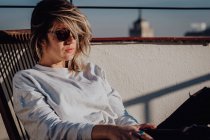 Jovem senhora elegante em casual wear e óculos de sol sentado na cadeira no telhado e céu azul com topo de torre em Madrid, Espanha — Fotografia de Stock