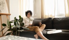 Jovem mulher feliz atraente sentado no sofá e usando laptop perto do conjunto de tintas em folhas na sala de luz — Fotografia de Stock