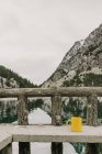 Taza amarilla en el banco cerca de la increíble vista de la superficie del agua entre altas montañas con árboles en la nieve y el cielo nublado en los Pirineos - foto de stock